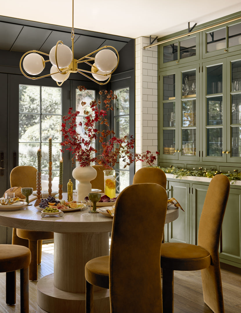 Velvet high back dining chairs surround a round dining table set with dinnerware, linens and decor for holiday hosting.