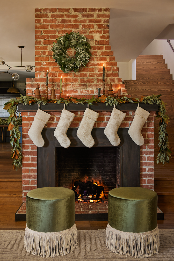 Two velvet green fringe ottomans sit in front of a fireplace and mantel decorated for the holidays with stockings, fresh handmade wreath and garland and decorative wooden trees and candlesticks.