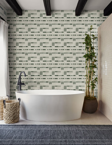 | The Artemis green and white dash-and-bar motif patterned wallpaper is in a bathroom with a white oval bathtub, a slate gray runner rug, and a natural tiled wall