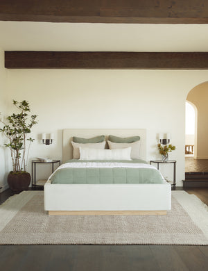 The Joelle rust rug lays in a bedroom under a natural linen frame bed that is in between two half-circle shaped night stands
