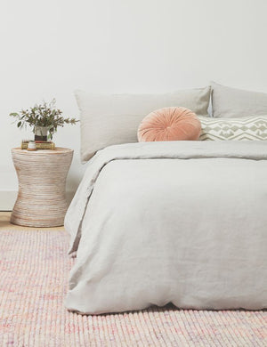 The European Flax Linen smoke gray Duvet Set by Cultiver lays on a bed in a bedroom with a pink woven rug, a pink velvet disc throw pillow, and a wooden ringed nightstand