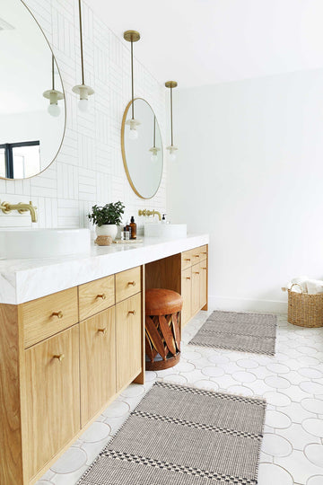 #size::1-9--x-2-10- #size::2--x-5- | Two Masha gray and black geometric machine washable mats lay in a bathroom with circular golden mirrors and marble countertops