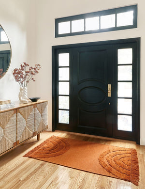 The Arches rust orange rug lays in an entryway with a large black front door framed by mirrors and a woven sideboard