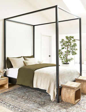 The European Flax Linen olive green Duvet Set by Cultiver lays on a black canopy bed in a bedroom with cream waffle linens, a persian rug, and natural wooden ottomans and nightstands
