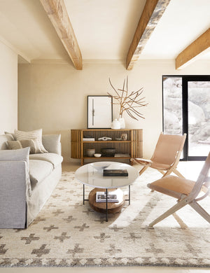 The Acoma cream and tan plus-sign patterned Moroccan area rug with diamond border lays in a neutral living room with a light gray slipcovered sofa and two leather folding chairs.
