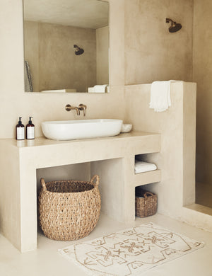 The Evet neutral geometric wool floor mat lays in a stone bathroom in front of a stone vanity with basin sink.