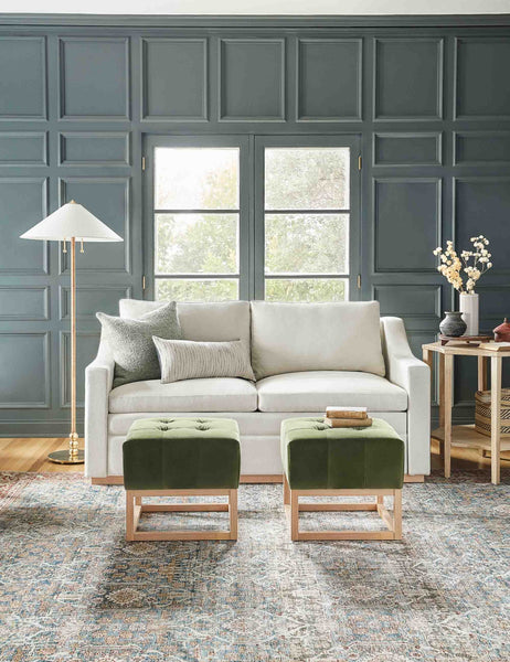 | The Zora floor lamp with polished base and brass knob detailing sits in a living room with a gray accented wall, a gray lining sofa, and two green velvet ottomans 