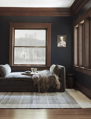 The Joelle black rug lays in a dark room under a gray velvet daybed with a plush throw atop it