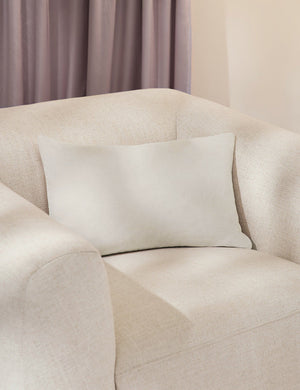 Charlotte oyster white lumbar velvet pillow sits on a white accent chair with a purple curtain in the background
