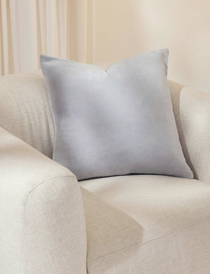 Charlotte ice blue square velvet pillow sits on a white accent chair with a white curtain in the background