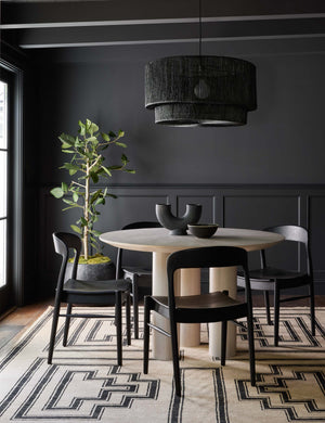 The Ida black teak wood dining chair sits in a dark living room with black walls surrounding a light wood circular dining table underneath a black jute chandelier.
