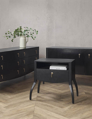 The Anabella black wood nightstand sits in a studio with books sitting in its open shelf and is next to the Anabella dresser