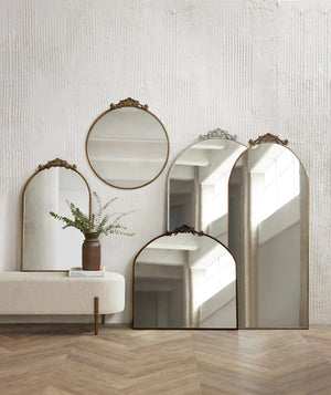 The Tulca arched oil rubbed bronze mirror with flat bottom edge and traditional scroll detailing sits on a chevron hardwood floor surrounded by other Tulca mirrors in their circular, narrow, and floor sizes.