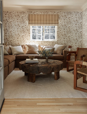 The beachwood rug lays in an enclosed living room with botanical wallpaper under a brown sectional and round coffee table