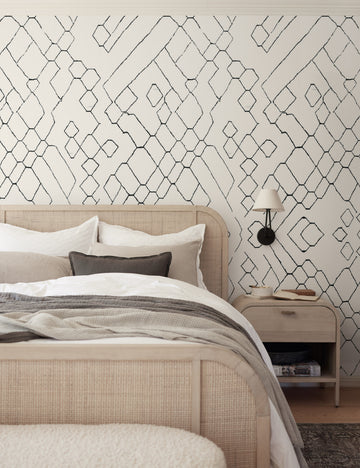 #color::black-and-ivory | The Moroccan black and ivory Wallpaper Mural by Sarah Sherman Samuel is in a bedroom with a woven cane bed, a light wood nightstand, and a black double armed sconce.