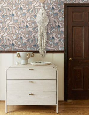 The mosaic wallpaper is in a room with a whitewashed three drawer dresser, a sculptural white vase, and a white woven wall hanging