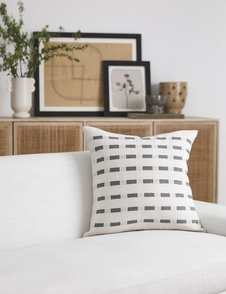 #color::pumice | Bertu pumice gray pillow with a woven dash pattern by Bolé Road Textiles sits on a white sofa with a rattan cane sideboard in the background