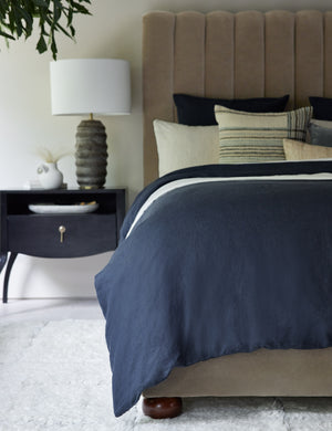 The European Flax Linen navy blue Duvet Set by Cultiver lays on a gray velvet framed bed in a bedroom with a black nightstand, a ribbed lamp, and a white plush rug
