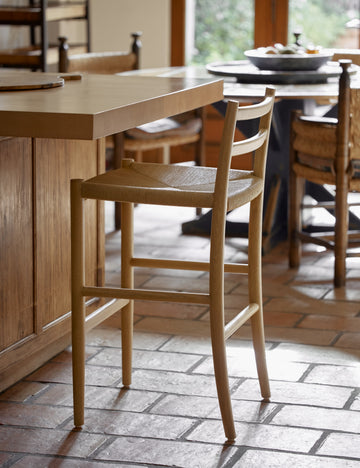 #color::natural-oak | Side view of the Nicholson slim natural oak wood frame and woven seat counter stool at a kitchen counter.