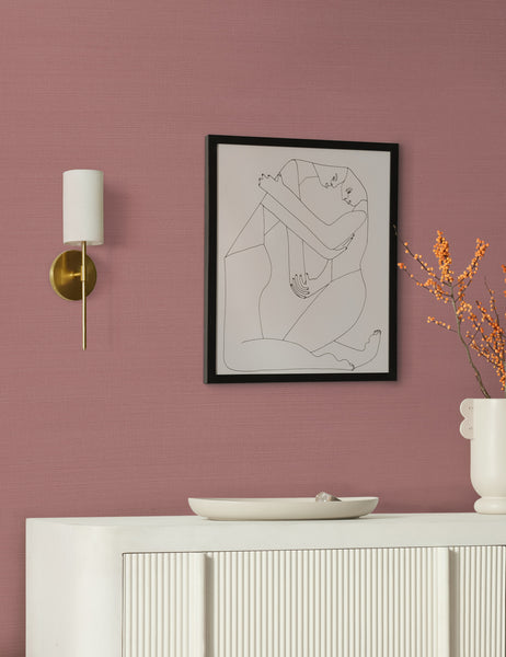 #color::aged-brass | The Jensine aged brass slim sconce with cylindrical shade is mounted on a pink wall to the right of a painting and above a white ribbed side table 