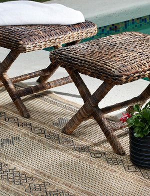 The Ember brown patterned flatweave indoor and outdoor rug lays beside a pool with two jute woven stools and a blue vase sitting atop it