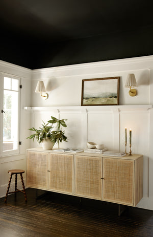 The Hannah natural mango wood sideboard with cane doors sits against a white accent wall underneath two white sconces and a landscape painting with a stack of books and candlesticks sitting atop it.