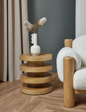 The Pentwater Four-Tiered Natural Round Side Table sits next to a boucle accent chair with a sculptural white vase