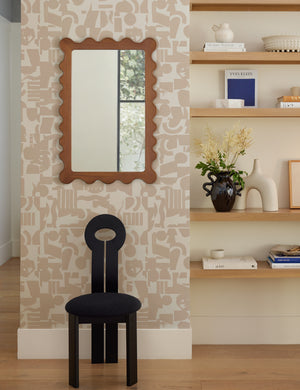 The Ripple mirror hangs on a patterned wall above a black sculptural chair next to a shelf with decor