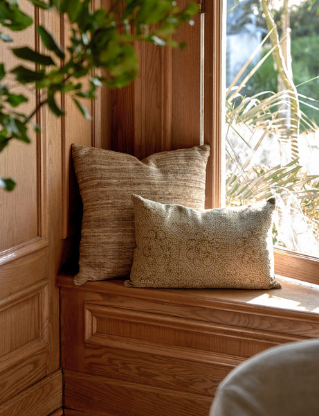 | The Bryce natural-toned silk square pillow sits in the corner of a room with accented wooden walls next to a window
