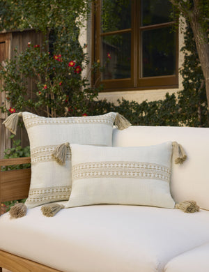 Marchesa natural and khaki indoor and outdoor pillow with tasseled corners sits on a natural-toned sofa next to a house with vines