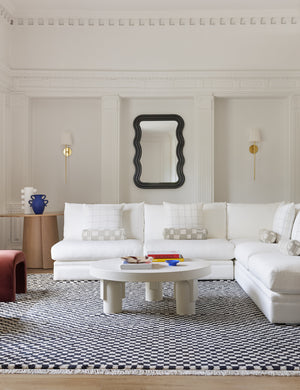 The Wendolyn wavy thick-framed black wall mirror hangs on a white paneled living room wall behind a white sectional sofa, black and white checkerboard rug and white round coffee table.