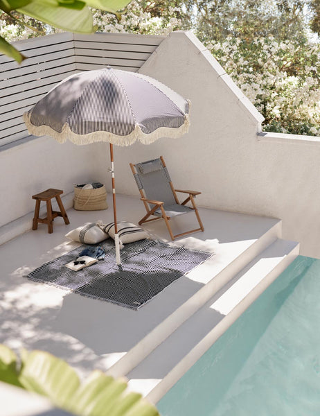 #color::navy-stripe | The Navy and white striped cotton beach blanket by business and pleasure co lays beside a pool with a striped beach chair and umbrella