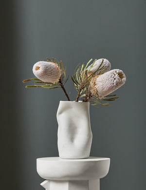 The Caverns white sculptural vase by Salamat Ceramics is filled with flowers and sits atop a white pedestal with a blue painted wall in the background