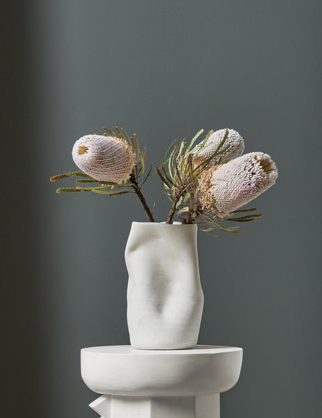 | The Caverns white sculptural vase by Salamat Ceramics is filled with flowers and sits atop a white pedestal with a blue painted wall in the background