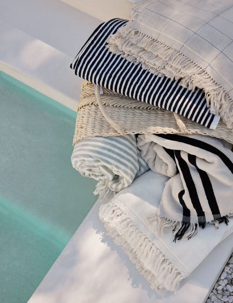 #color::pop-check #color::navy-stripe #color::sage-stripe #color::antique-white #color::black-stripe #color::antique-white | Beach Towel by Business & Pleasure Co in all its colors sit in a stack next to a pool