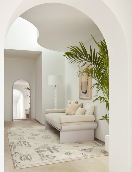 #color::silver | The Shashenka silver arched floor mirror sits in a sculptural space with a cream cushioned bench, a plush patterend rug, and a white lamp with a ribbed base