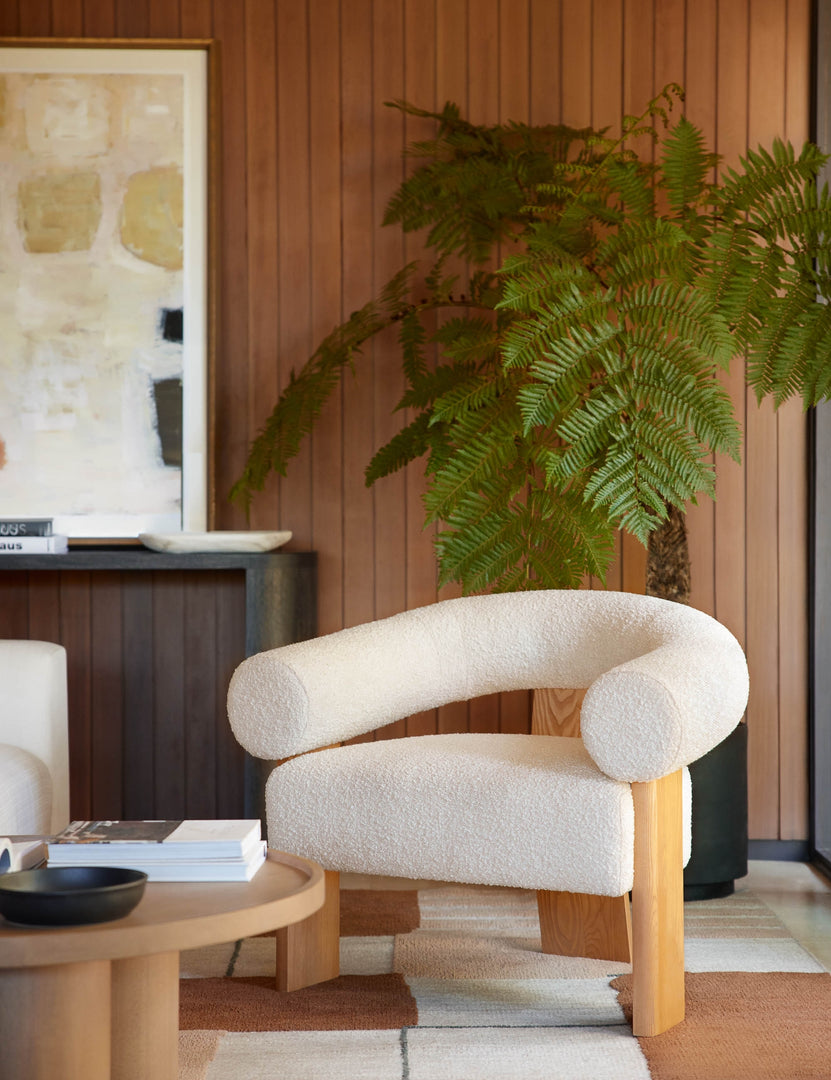 #color::cream | The Celeste honey wood accent chair with wishbone frame sits in a retro living room with wood paneled walls, a black planter, and a round wooden coffee table.