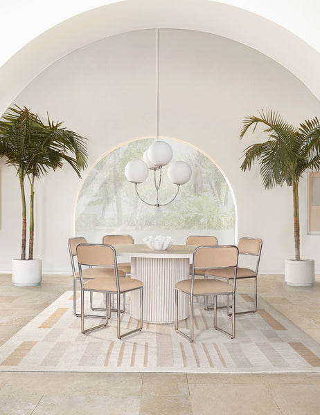 #color::white-wash | The Rutherford white-washed acacia wood round dining table with pedestal base sits in an airy dining room surrounded by six light pink dining chairs atop a natural patterned rug with two palm trees and a large archway behind it.