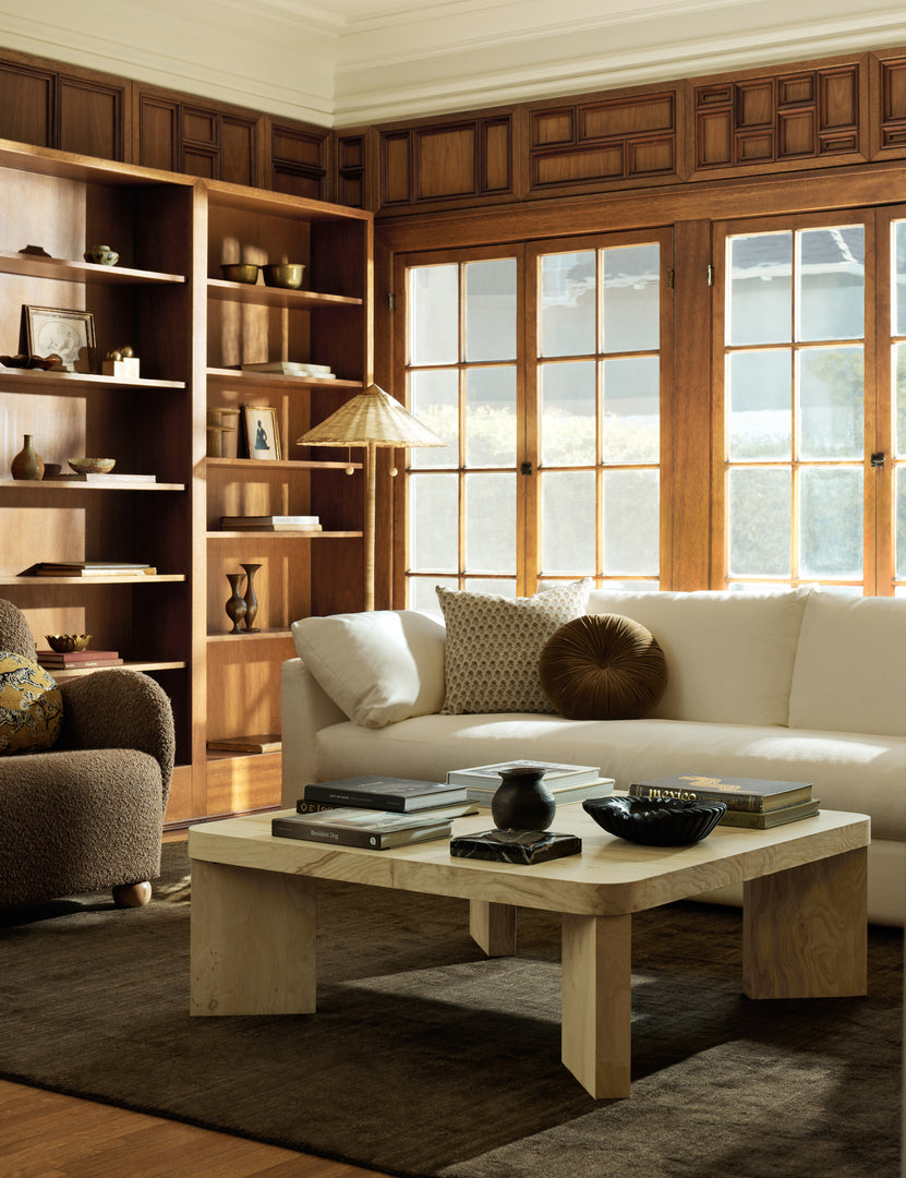 In a lounge a white sofa and brown boucle upholstered accent chair are paired with a square burl wood coffee table.