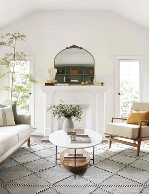 The Tulca arched gold mirror with flat bottom edge and traditional scroll detailing sits atop a fireplace in a bright living room with a natural sofa and accent chair and a black and white patterned rug.