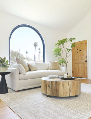 The Boni round coffee table sits in a living room with a large arched window, an ivory rug, and an ivory sofa