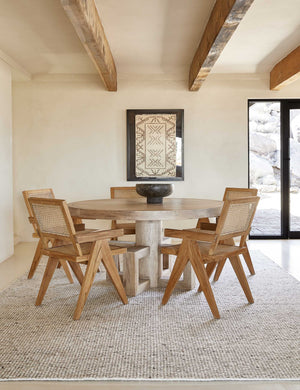 The Taos neutral light brown wool blend area rug lays under a round light wood dining table surrounded by five wood and cane arm chairs.