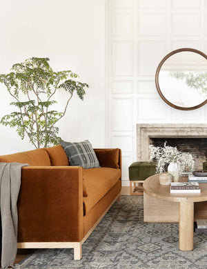  Cognac Velvet Hollingworth Sofa sits in a living room with a dark-toned patterned rug, a green velvet ottoman, and an oval coffee table