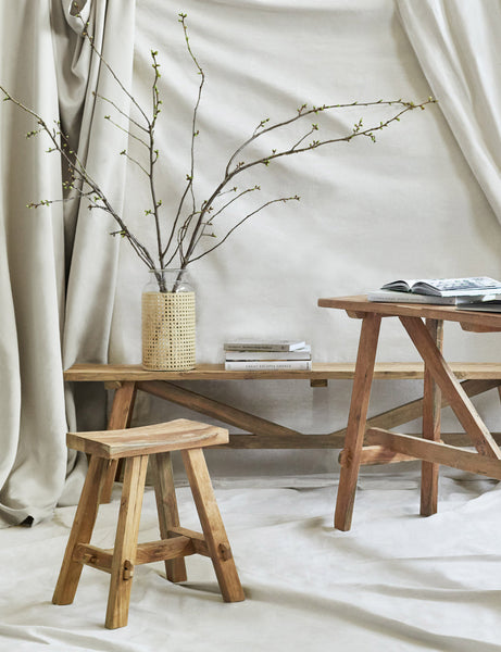 | The Arlene craftsman-style antiqued teak wood bench, along with the matching Arlene stool and console table are displayed against a wall draped in light tan fabric.