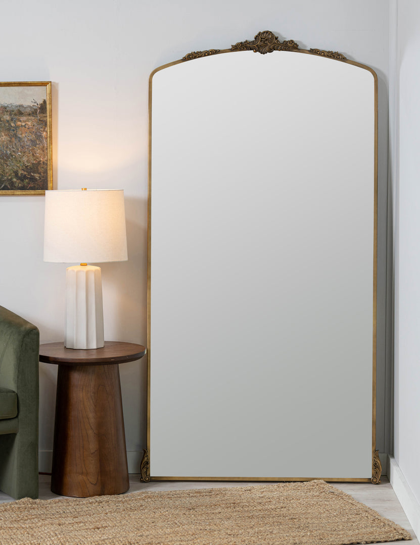 #color::gold | Casserly ornate gold full length mirror leaning against a wall next to a round side table with lamp.