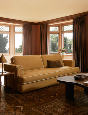  Golden ochre two-cushion sofa with contrast cream piping and clean track arms, styled with a leopard print pillow in a terracotta living room with floor-length mocha drapes