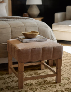  The Ambleside apricot linen ottoman sits at the edge of a bed with a book and a wooden bowl atop a patterned rug