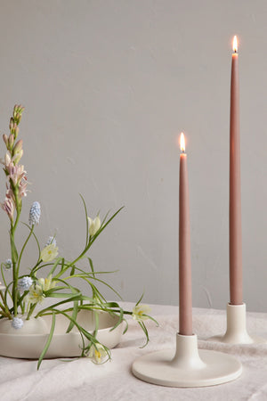 Two white sleek Ceramic Taper Candle Holders with dusty pink lit taper candles sit atop a table to the right of a vase filled with flowers