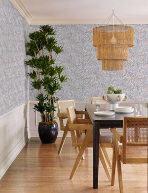 The Chavette three-tiered jute-wrapped chandelier hangs over a black dining table surrounded by light wood chairs with cane backs in a dininr room with blue abstract wallpaper.