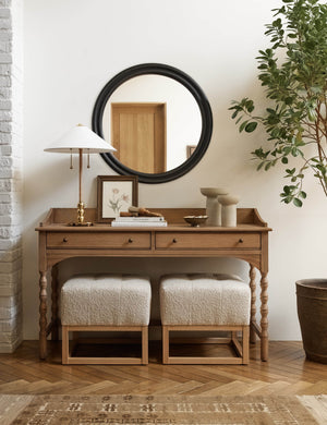  Two Taupe Boucle Grasmere Ottomans sit in a room beneath an antique desk, a table lamp, and a black framed circular mirror